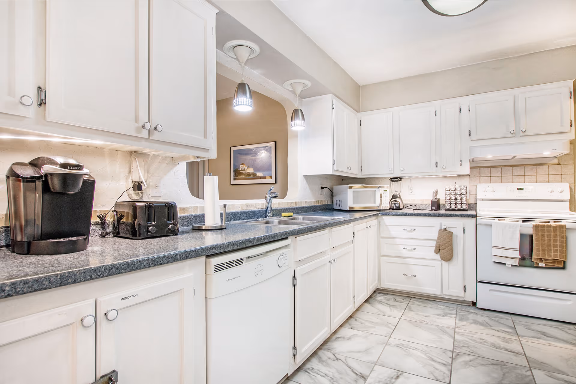 A bright kitchen with white cabinets and drawers, a gray countertop, and a tiled floor. The kitchen features a Keurig coffee maker, a toaster, a paper towel holder, a dishwasher, a microwave, a blender, a spice rack, and a white stove with oven mitts hanging on the handle. Two pendant lights hang over the sink area, and a framed picture is visible on the wall in the adjacent room.