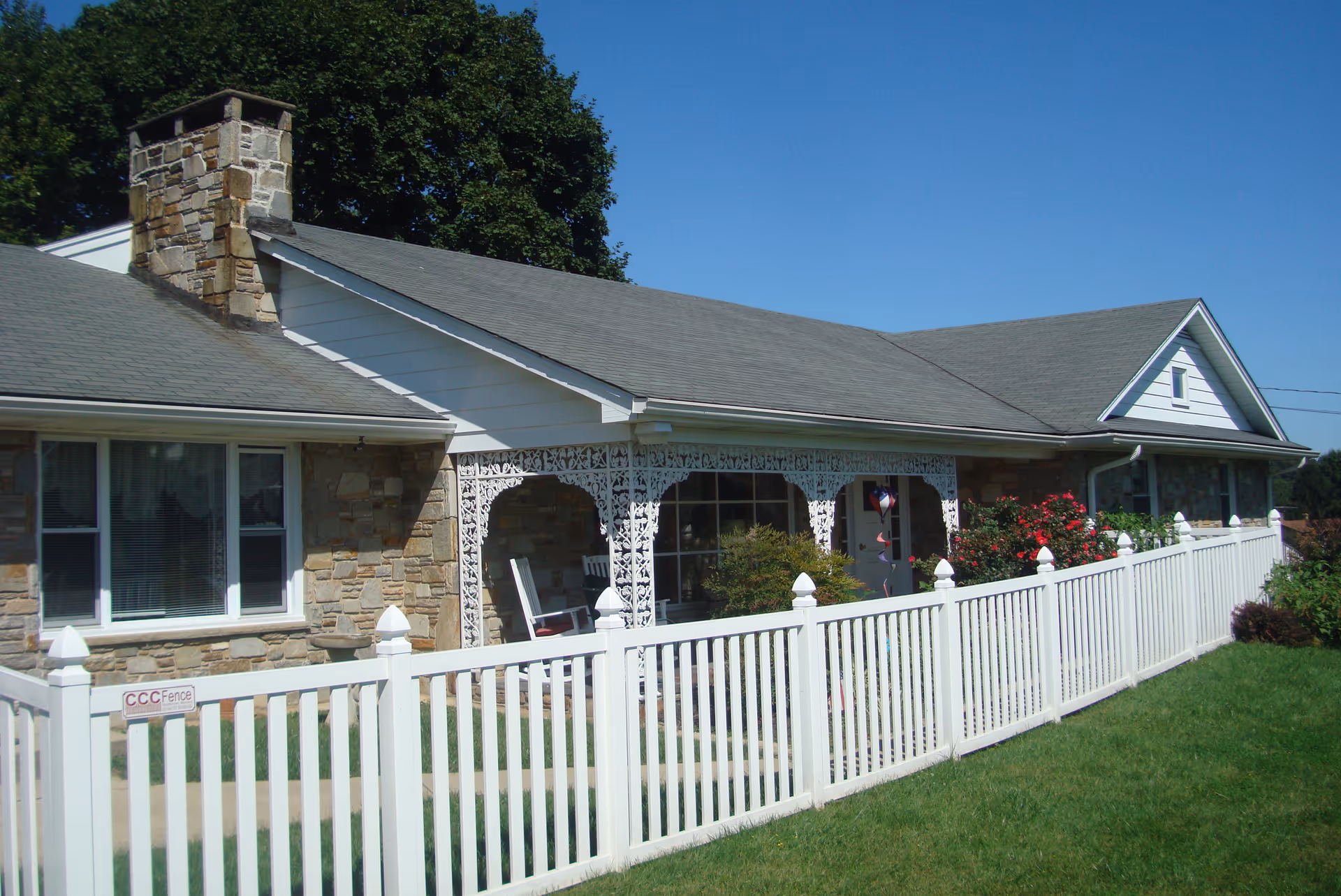 Front exterior of a single-story home with stone siding, a decorative covered porch, and a white picket fence under a clear blue sky.