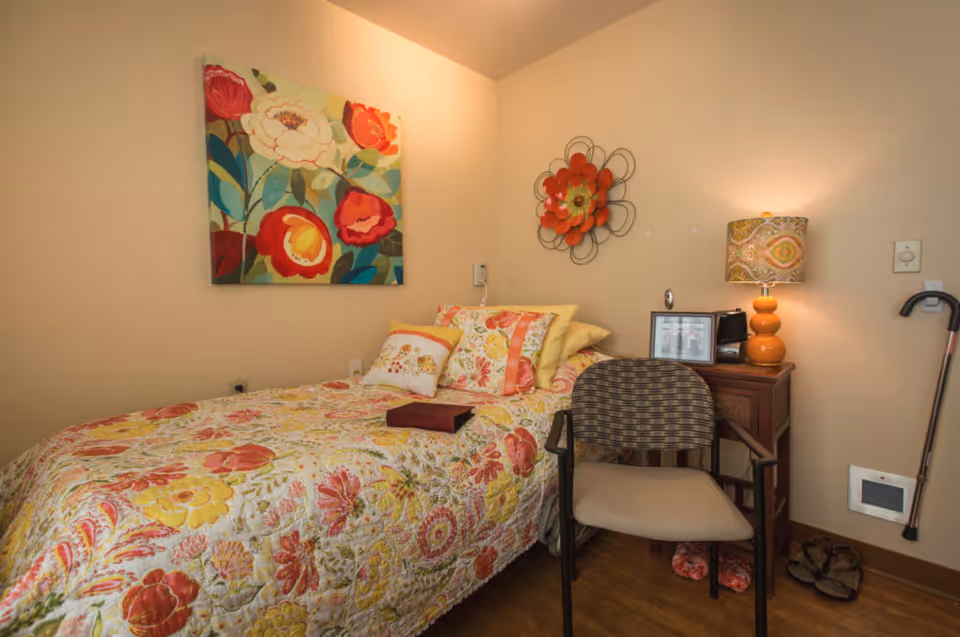 A cozy bedroom with a single bed covered in a colorful floral quilt and matching pillows. Above the bed is a vibrant floral painting and a decorative wall flower. Next to the bed is a wooden nightstand with a patterned lamp, framed photos, and a small clock. A cushioned chair is placed in front of the nightstand. On the floor near the nightstand are a pair of slippers and a walking cane hanging on the wall.
