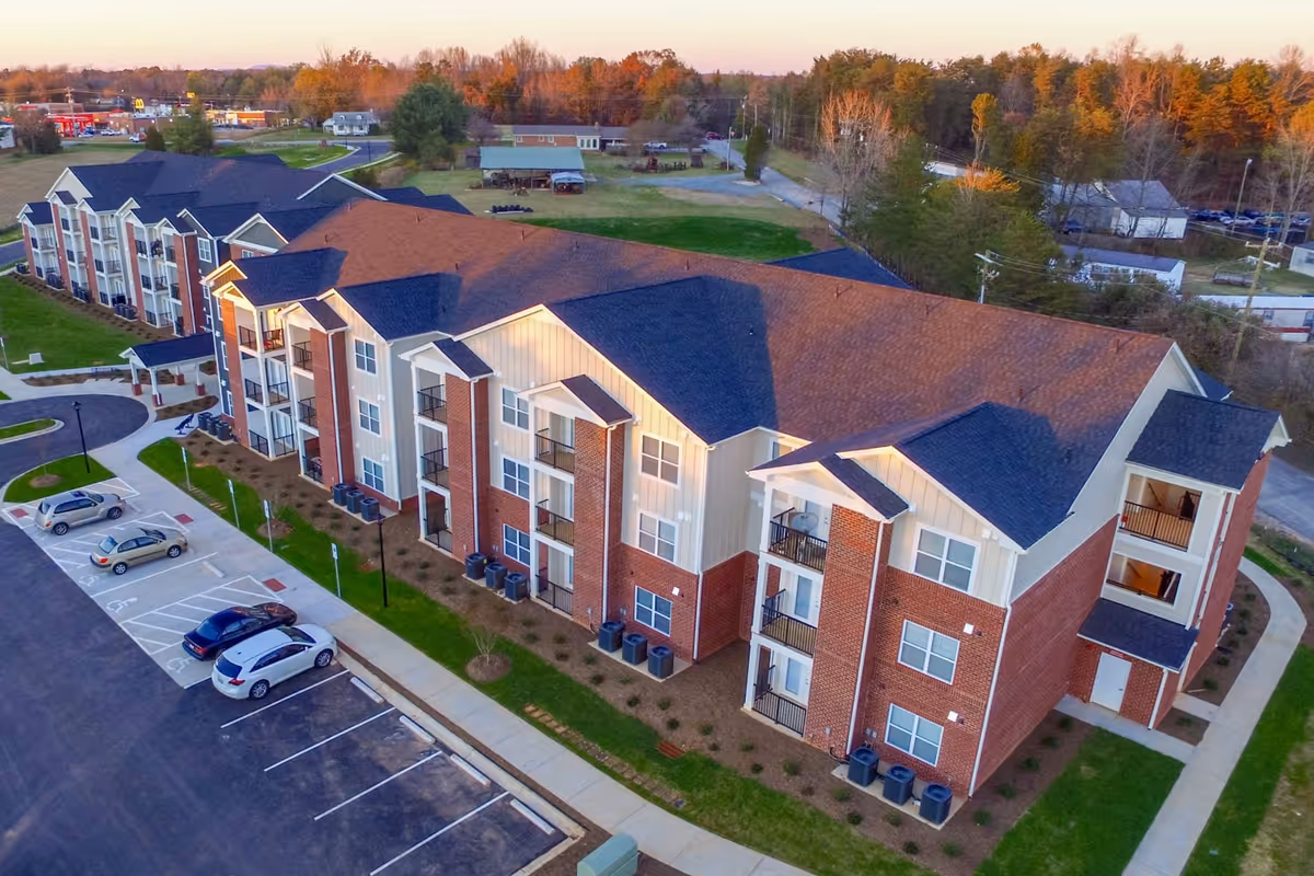 Aerial view of a multi-story brick and siding senior living building with balconies, walkways and a parking lot at sunset.