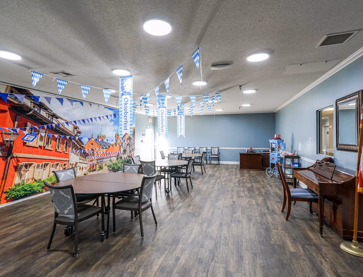 A spacious common room decorated with blue and white Oktoberfest banners and pennants hanging from the ceiling. The room has several tables and chairs arranged for seating, a piano with a chair, a popcorn machine, and a large mural on one wall depicting a European street scene with buildings and blue sky. The floor is wood-style, and the walls are painted light blue.