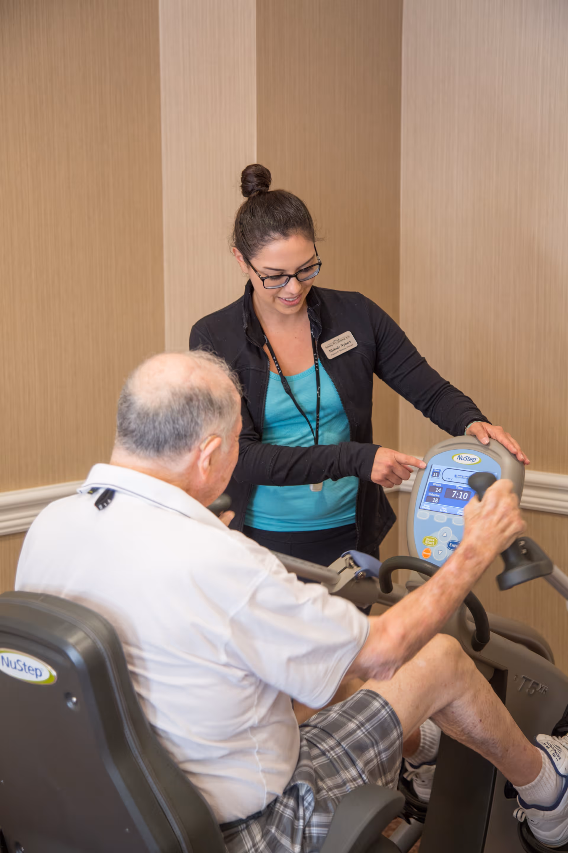 A female staff member with glasses and a name tag is assisting an elderly man using a NuStep exercise machine in an indoor setting with beige walls.