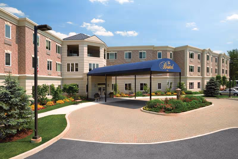Front exterior view of The Bristal Assisted Living at Armonk building with a covered entrance, landscaped garden beds, and a paved driveway under a blue sky with some clouds.