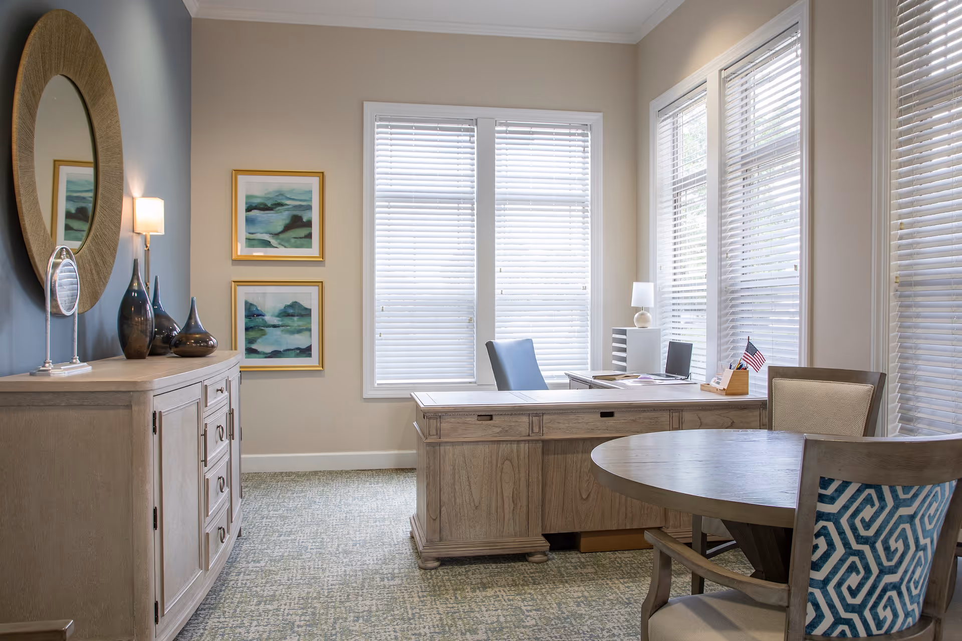 A bright office room with a wooden desk and a blue office chair positioned near large windows with white blinds. There is a round wooden table with two chairs, one with a blue patterned backrest. On the left side, a wooden cabinet holds decorative vases and a round mirror hangs above it. Two framed abstract paintings are on the wall next to the windows.