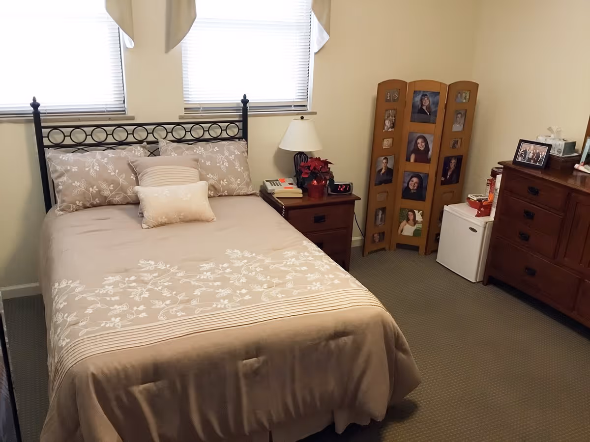 Cozy bedroom featuring a made double bed with patterned bedding, a nightstand and lamp, wooden dresser, small refrigerator, and a folding photo screen.