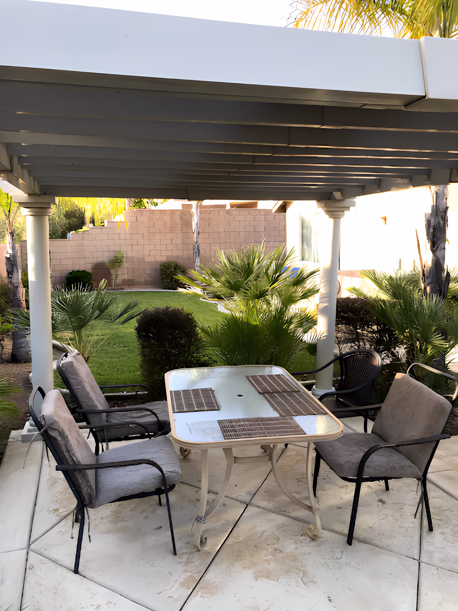 Outdoor patio area with a glass-top table and four cushioned chairs under a white pergola. The patio overlooks a green lawn with various bushes and palm plants, bordered by a brick wall.