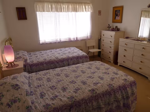 Bright bedroom with two twin beds in matching purple floral quilts, a window with curtains, a lamp on a nightstand, and white dressers.