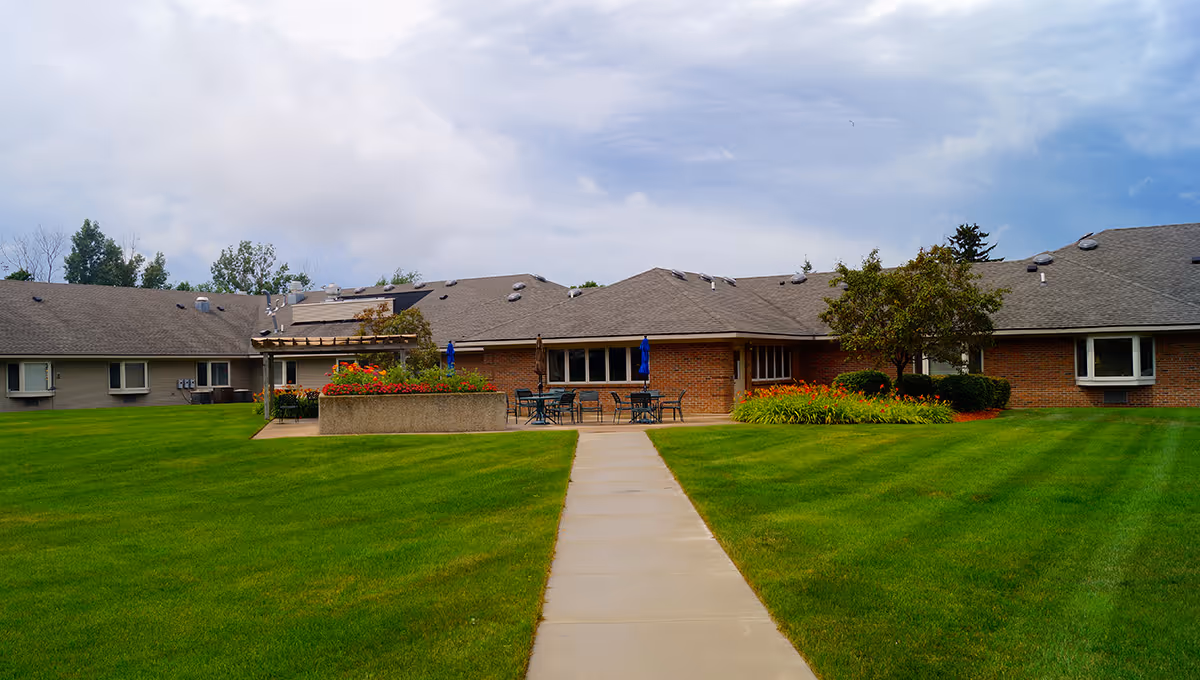 Wide exterior view of a single-story brick assisted living building with a central walkway, manicured lawn, and patio seating with umbrellas.