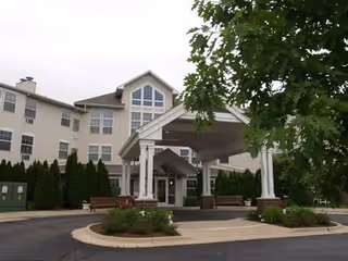 Front exterior view of a multi-story senior living facility named St Josephs Village with a covered entrance, benches, landscaped greenery, and a tree in the foreground.