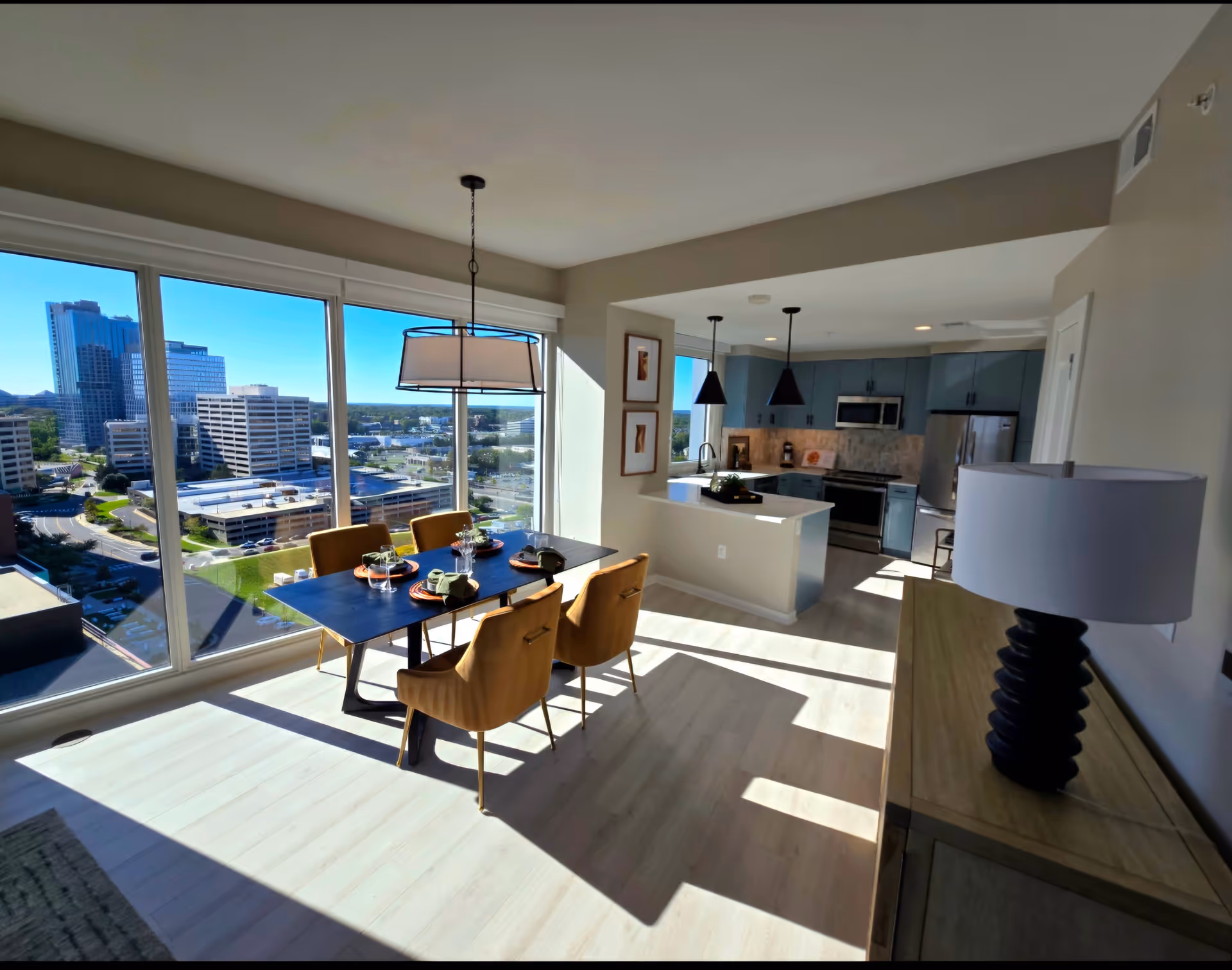 Sunlit dining area with a table and chairs next to floor-to-ceiling windows overlooking a cityscape, adjacent to a modern open kitchen.
