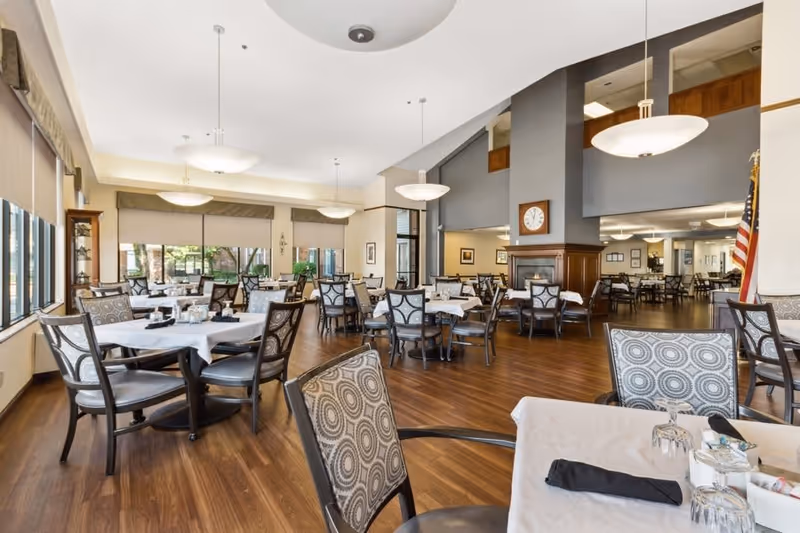 Spacious dining room in a senior living facility with multiple round tables set with white tablecloths and patterned chairs under large pendant lights.