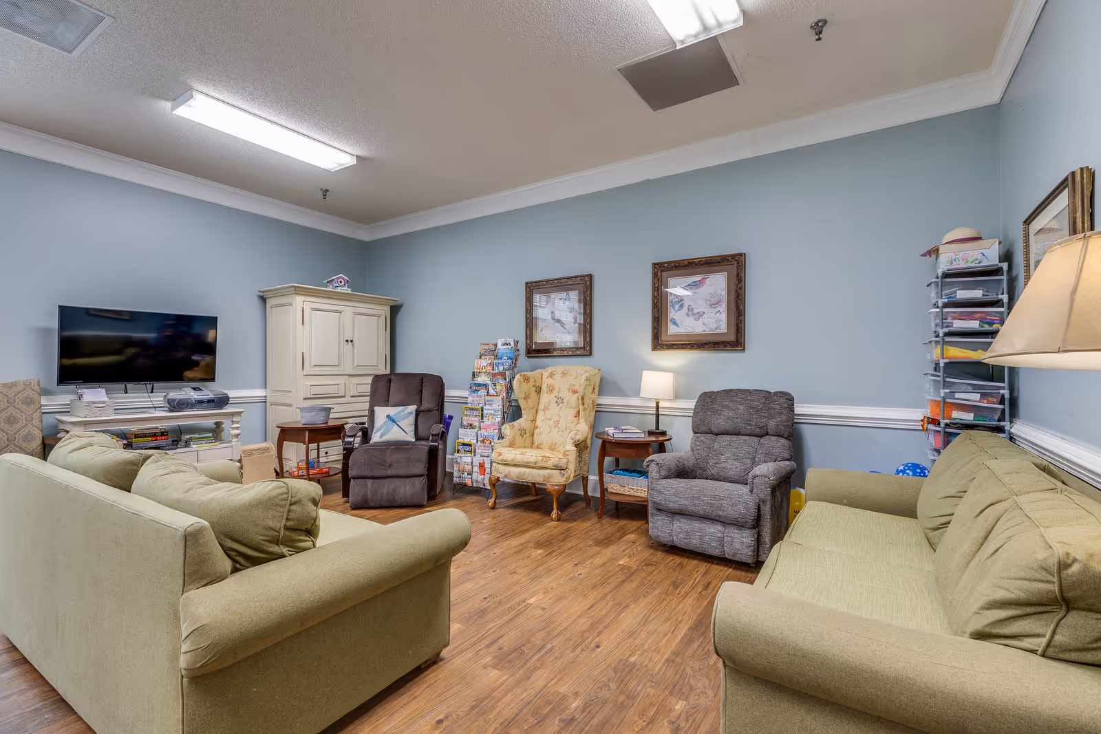 A cozy living room in an assisted living facility with two green sofas facing each other, two recliner chairs, a floral armchair, a TV on a stand, a white cabinet, a small side table with a lamp, and a rack filled with magazines and papers. The walls are painted light blue with white trim, and the floor is wooden.