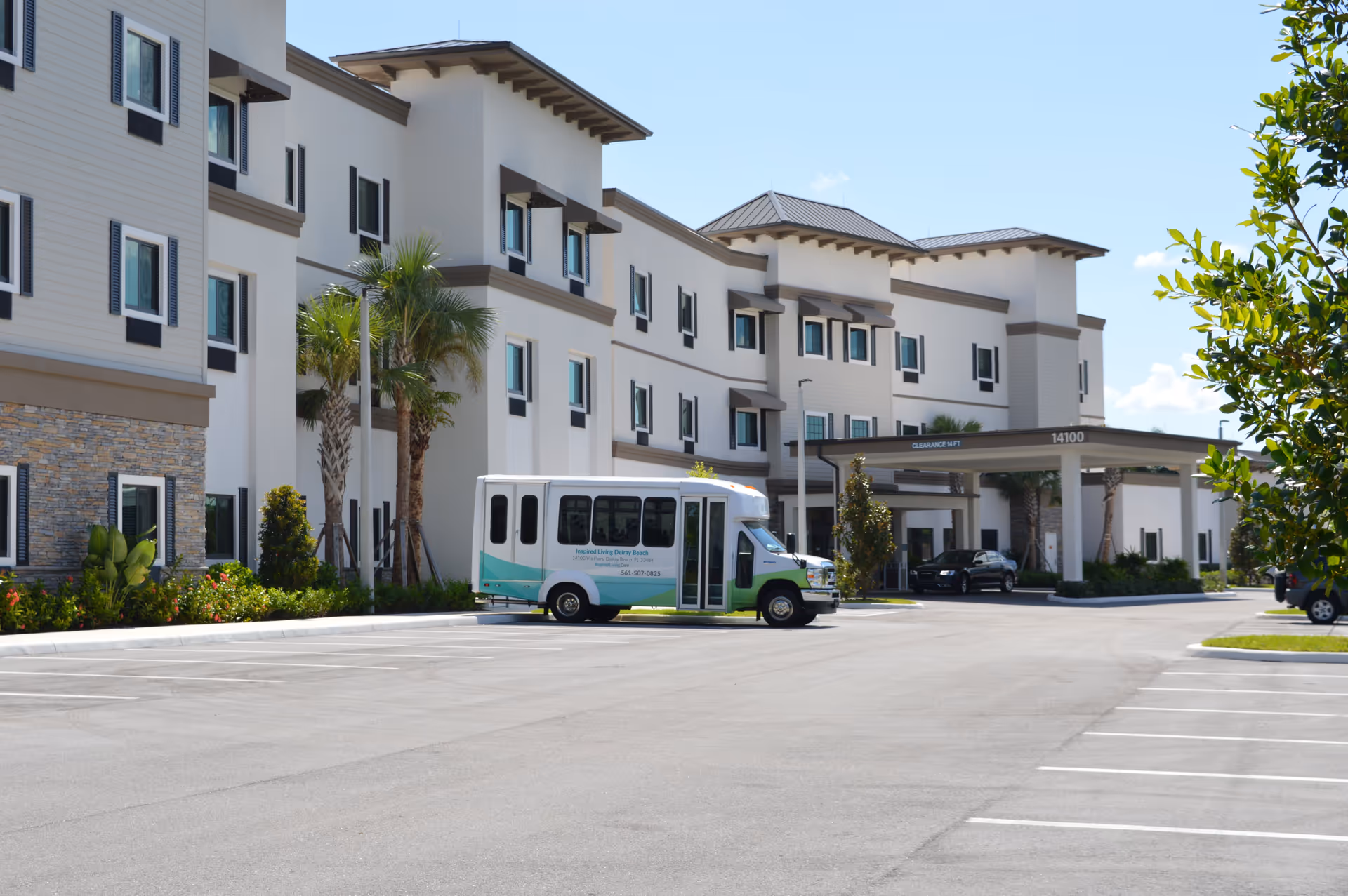Exterior view of a multi-story senior living facility with a covered entrance and a small shuttle bus parked in front. The building is light-colored with dark window shutters and palm trees along the side. The parking lot is mostly empty under a clear blue sky.