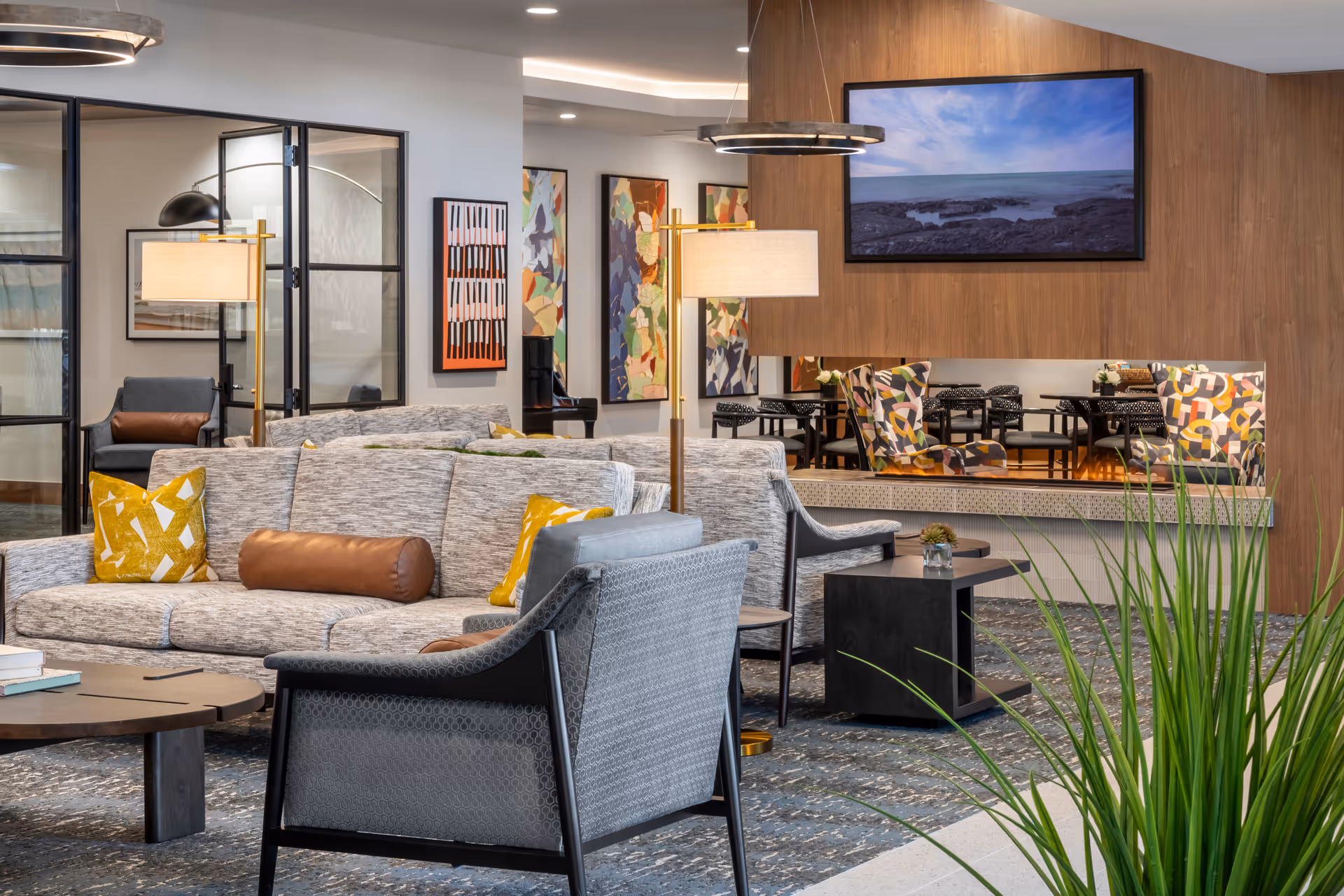A modern living room area with a gray sofa adorned with yellow and brown pillows, a gray armchair, a round wooden coffee table, and a floor lamp. The background features colorful abstract paintings, a wooden wall with a mounted TV displaying a coastal scene, and a large mirror reflecting patterned chairs and a dining area. There is also a green plant in the foreground.