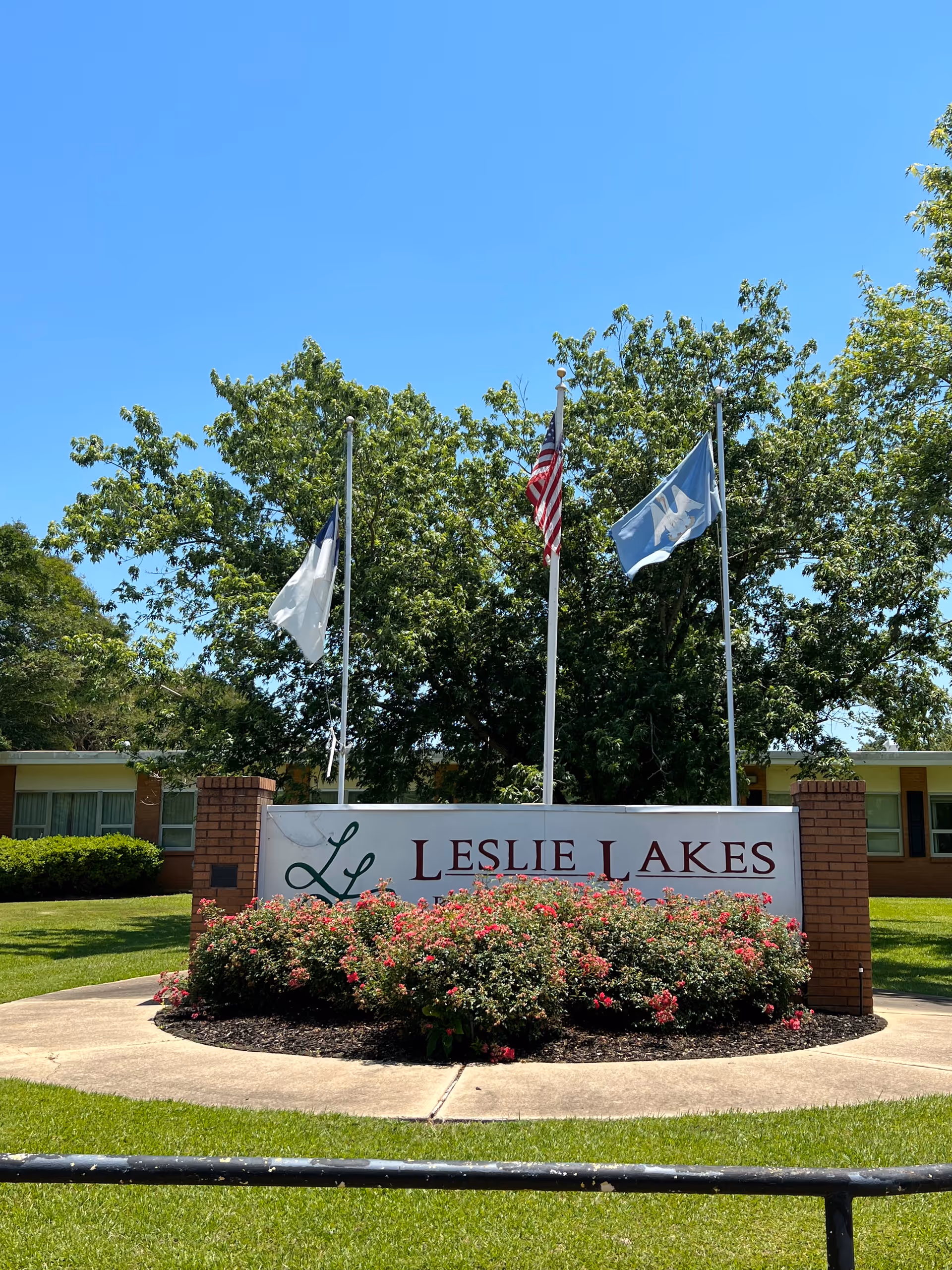 Entrance sign reading "Leslie Lakes" surrounded by flowering shrubs and flagpoles in front of the facility building.