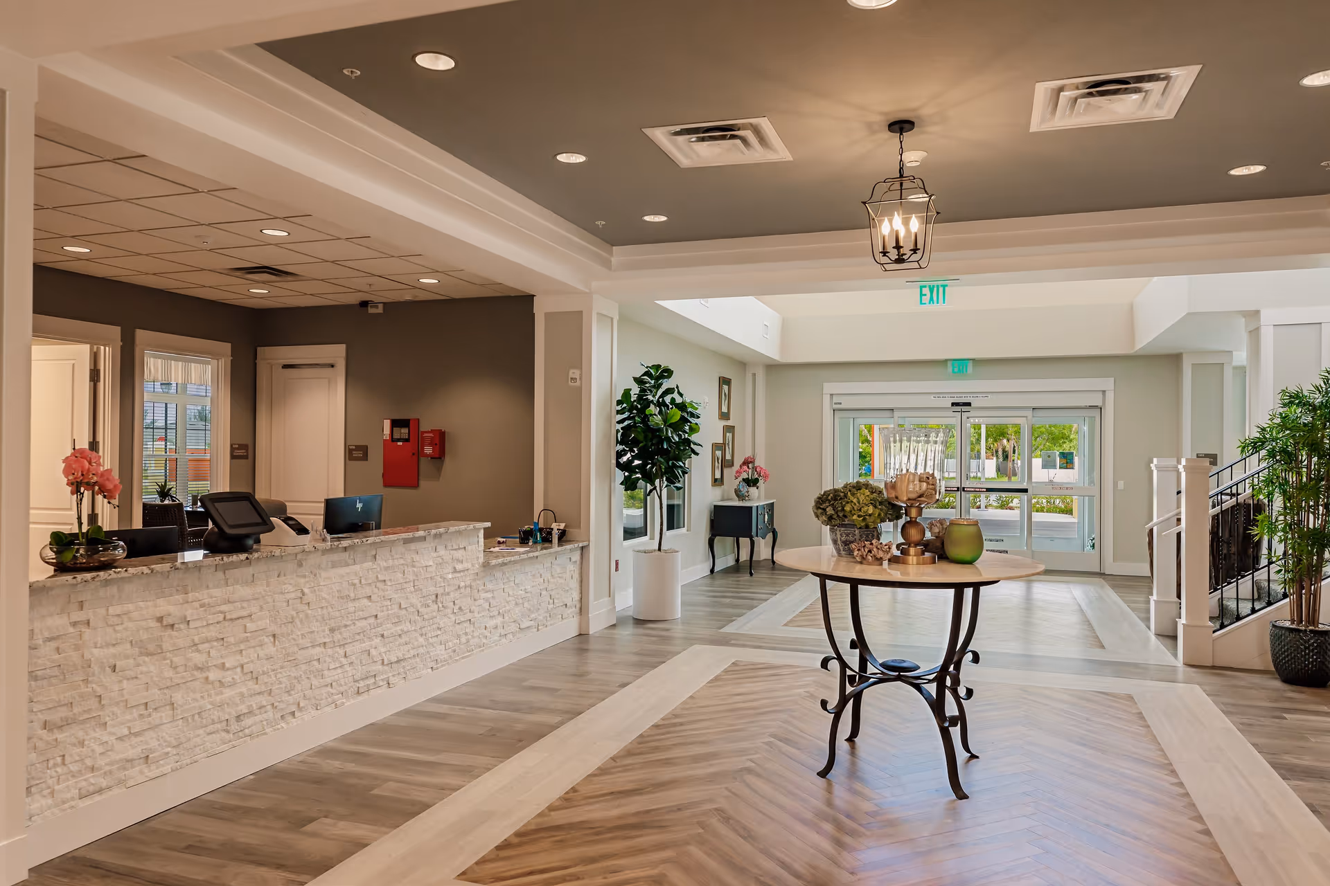 A spacious and well-lit reception area with a stone-textured front desk on the left, decorated with a pink orchid and office equipment. In the center, there is a round table with decorative items including a green vase and a candle holder. The floor has a wood pattern, and there are plants and framed pictures along the walls. Large glass doors at the back lead outside, and there is a staircase on the right side.