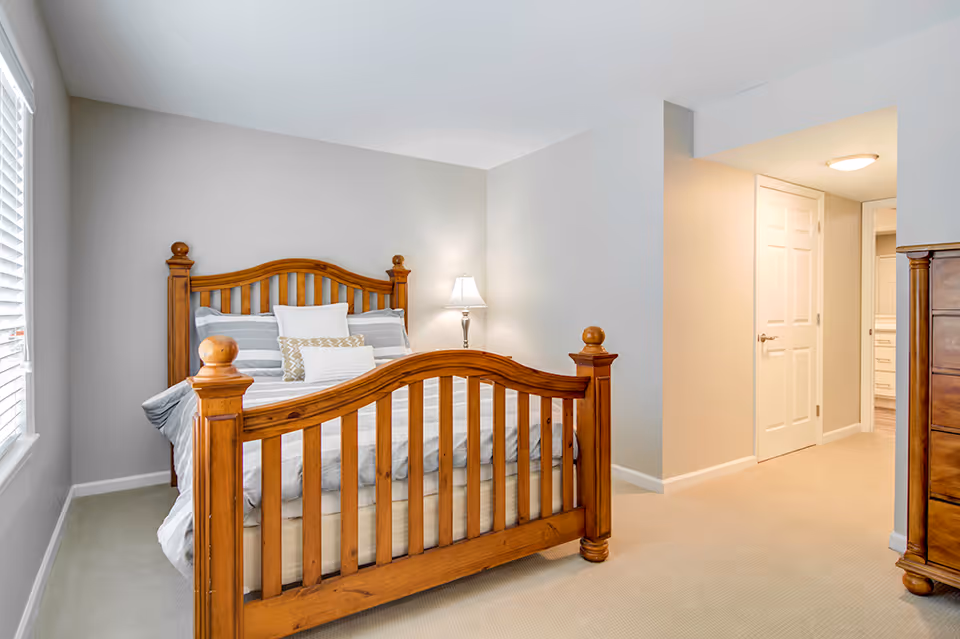 Bright bedroom with a wooden bed frame, striped bedding, bedside lamp, window with blinds, and an open doorway to a hallway.
