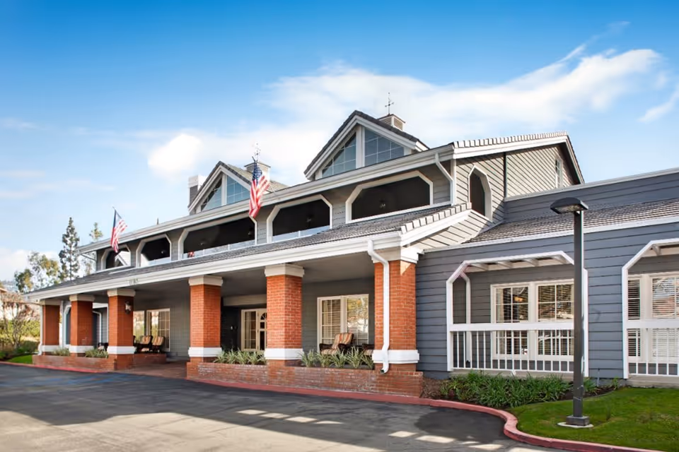 Exterior view of a senior living facility building with gray siding and red brick columns. The building has a covered porch with seating, American flags, and large windows with white trim. The sky is clear with a few clouds.