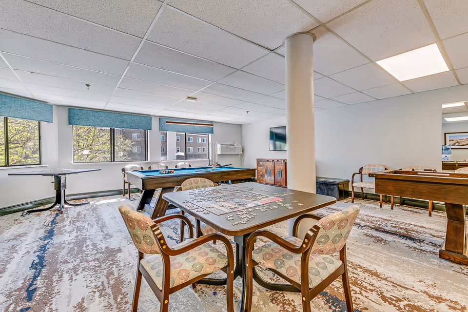 A bright recreational room with large windows letting in natural light. The room features a pool table, a table with a partially completed jigsaw puzzle, several cushioned chairs with patterned upholstery, and a TV mounted on the wall. The carpet has a modern abstract design, and there is a white column near the center of the room.