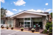 Front exterior view of Potomac Valley Rehabilitation and Healthcare Center building with a sloped roof, large glass entrance doors, stone pillars, potted plants, and a clear blue sky with some clouds.
