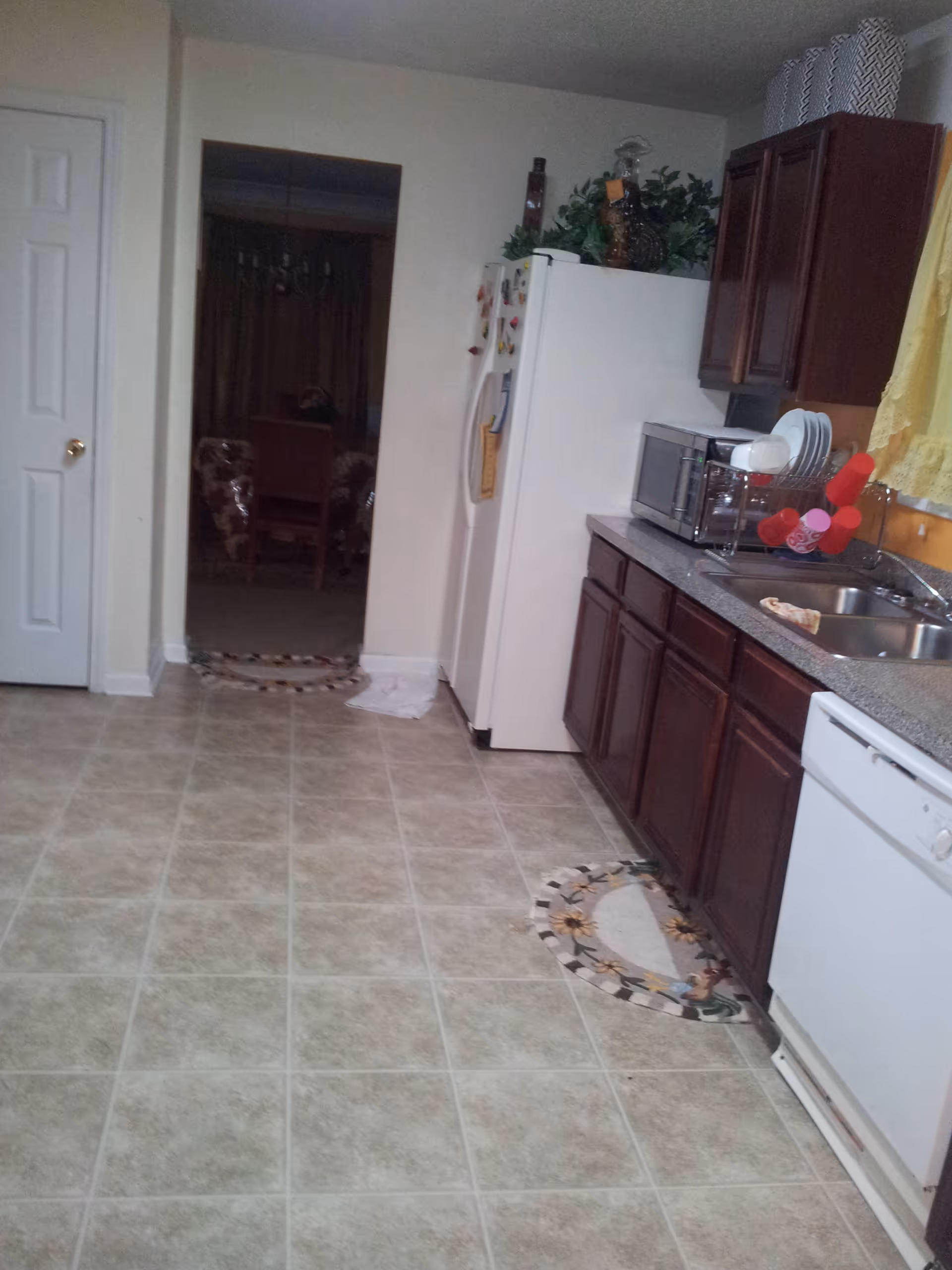 Interior view of a kitchen with tiled floor, dark wooden cabinets, a white refrigerator, a microwave, a double sink, and a dishwasher. There are decorative mats on the floor and a yellow curtain above the sink. A doorway leads to a dining area with a table and chairs.