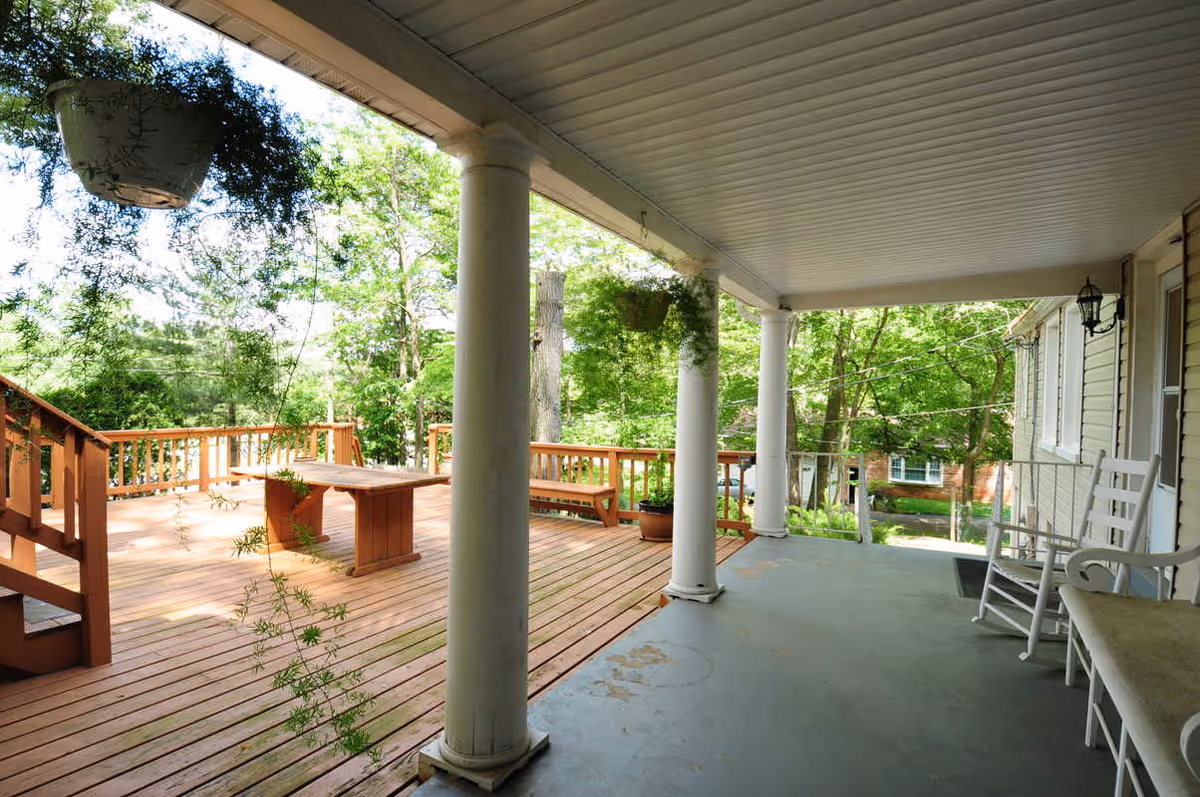 A covered porch area with white columns and rocking chairs overlooking a wooden deck with a table and benches, surrounded by green trees and plants.