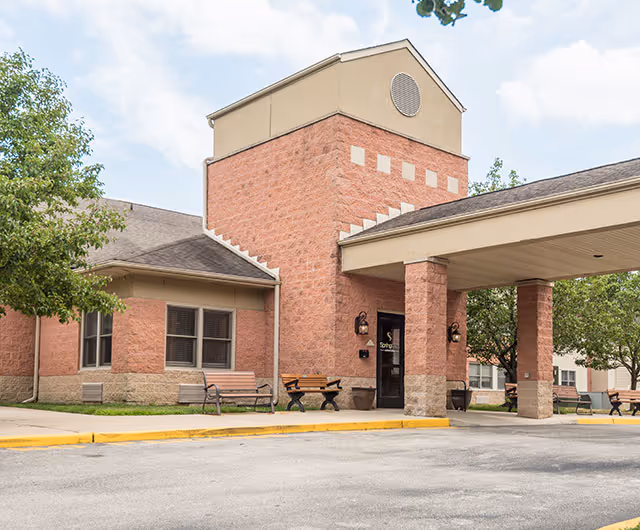 Front entrance of a brick senior living facility with a covered porte-cochere, tower feature, and outdoor benches.