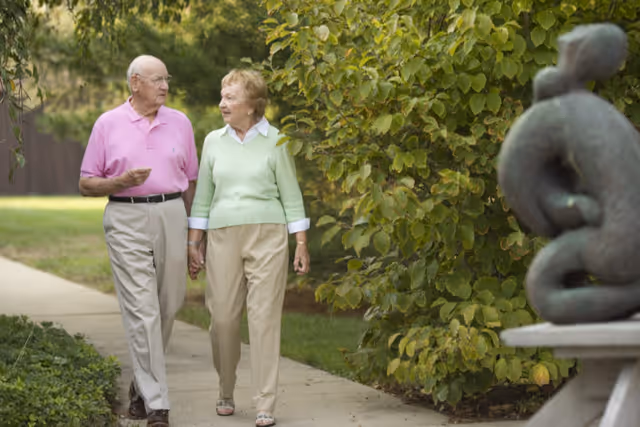 Two people holding hands and walking along a garden path beside leafy shrubs with a sculpture visible in the foreground.