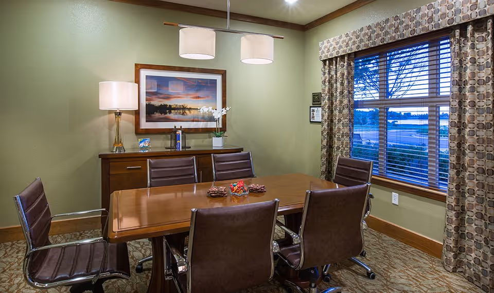 A formal meeting/dining room with a wooden table surrounded by six brown leather chairs, framed artwork and a lamp against a green wall, and a window with patterned curtains showing an outdoor view.