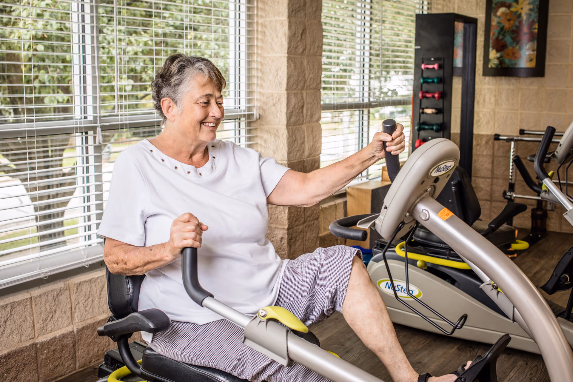 An elderly woman exercising on a recumbent stationary bike in a fitness room with large windows and various exercise equipment in the background.