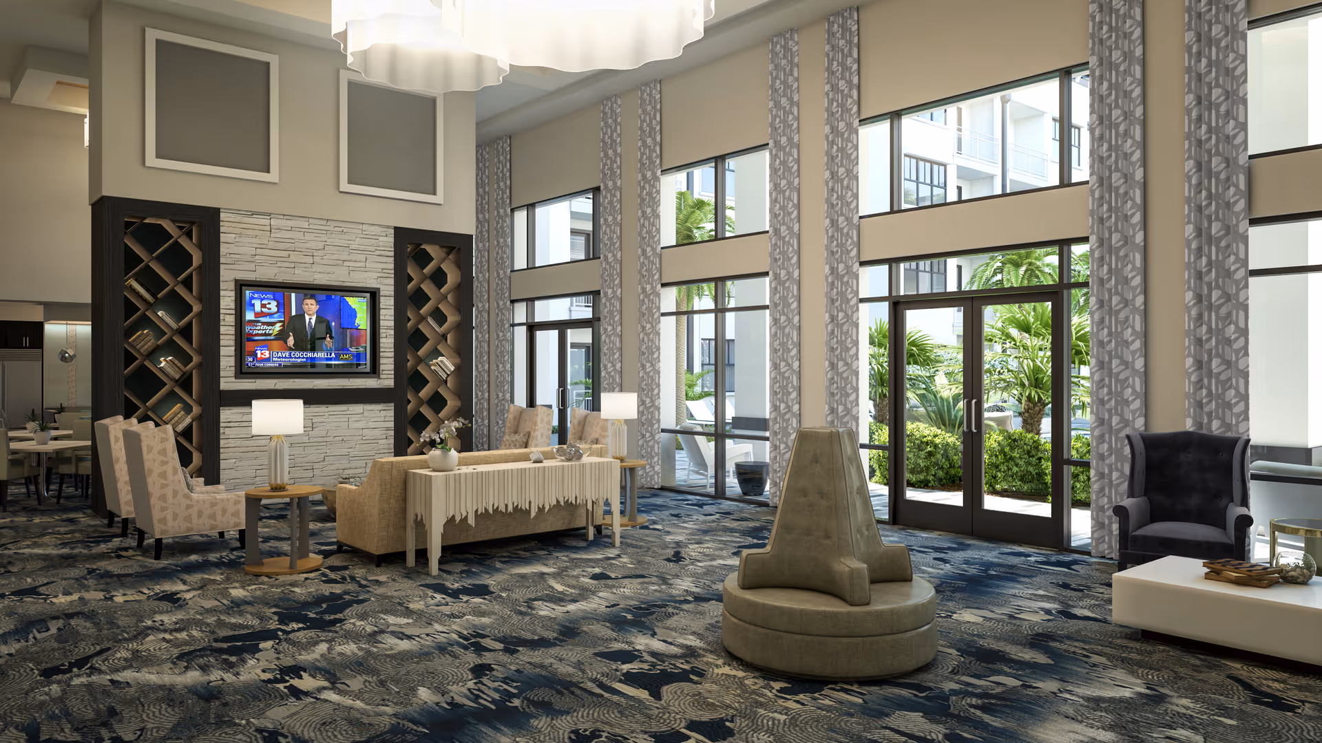 A spacious and well-lit living room area with large windows and glass doors that open to an outdoor garden. The room features a patterned blue and beige carpet, multiple seating options including beige armchairs, a central round cushioned seat, and a dark upholstered armchair. A flat-screen TV is mounted on a stone accent wall with built-in shelves on either side holding books. There are two table lamps on side tables and decorative elements on a console table behind a sofa.