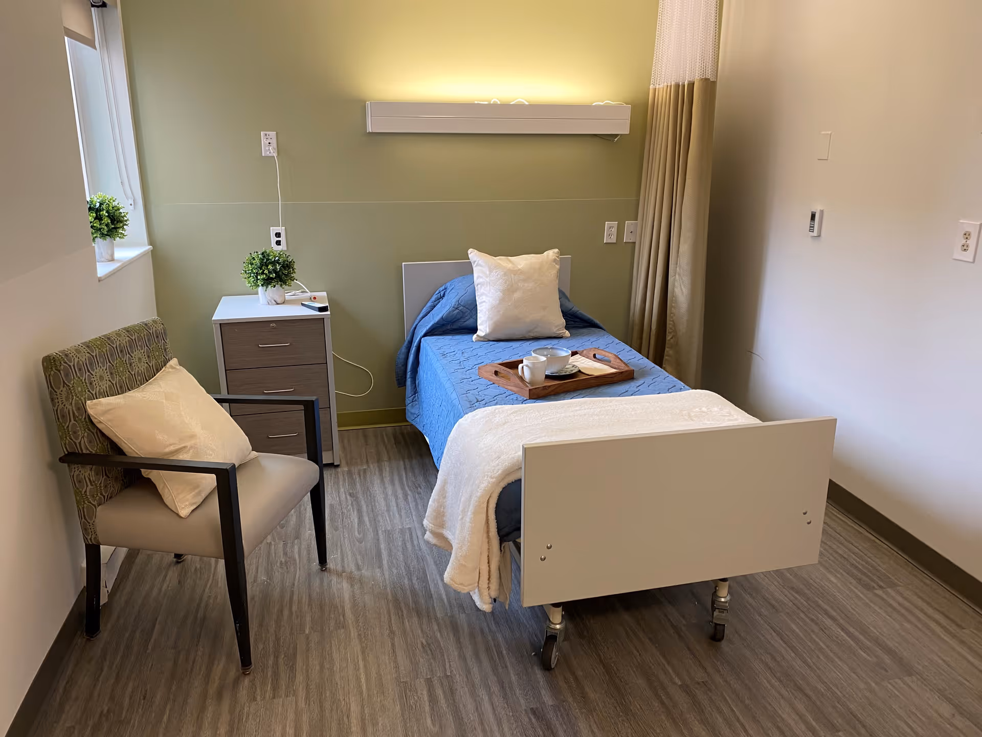 A small, neatly arranged bedroom in a healthcare facility featuring a single bed with blue bedding and a white pillow, a wooden tray with a cup and bowl on the bed, a bedside table with a small plant and remote control, a cushioned chair with a pillow, and a window with a potted plant on the sill. The room has light green and white walls and wood-patterned flooring.