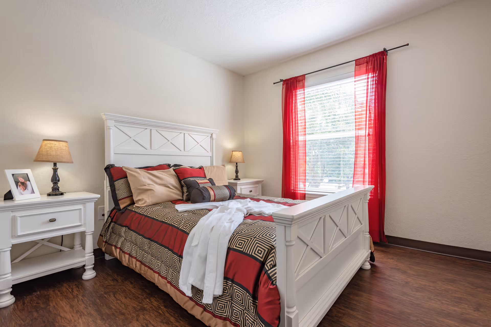 Well-lit bedroom with a white wooden bed dressed in red and gold patterned bedding, matching white nightstands and lamps, and a window with red curtains.