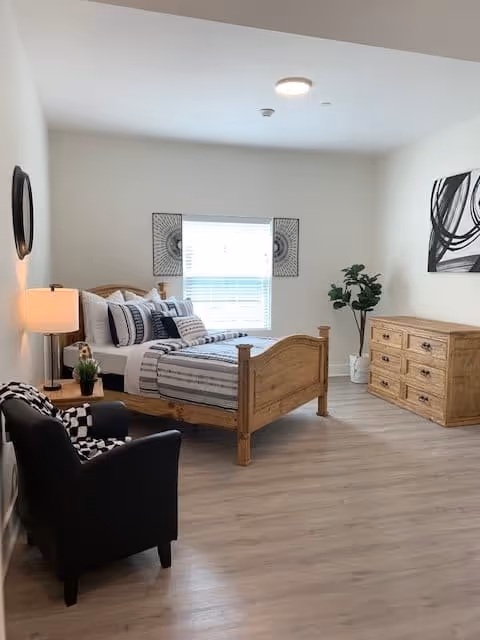 A bright bedroom with a wooden bed frame made with striped bedding and multiple pillows. There is a black armchair with a patterned blanket draped over it, a wooden dresser with six drawers, a potted plant in the corner, and two pieces of wall art. A window with blinds lets in natural light.