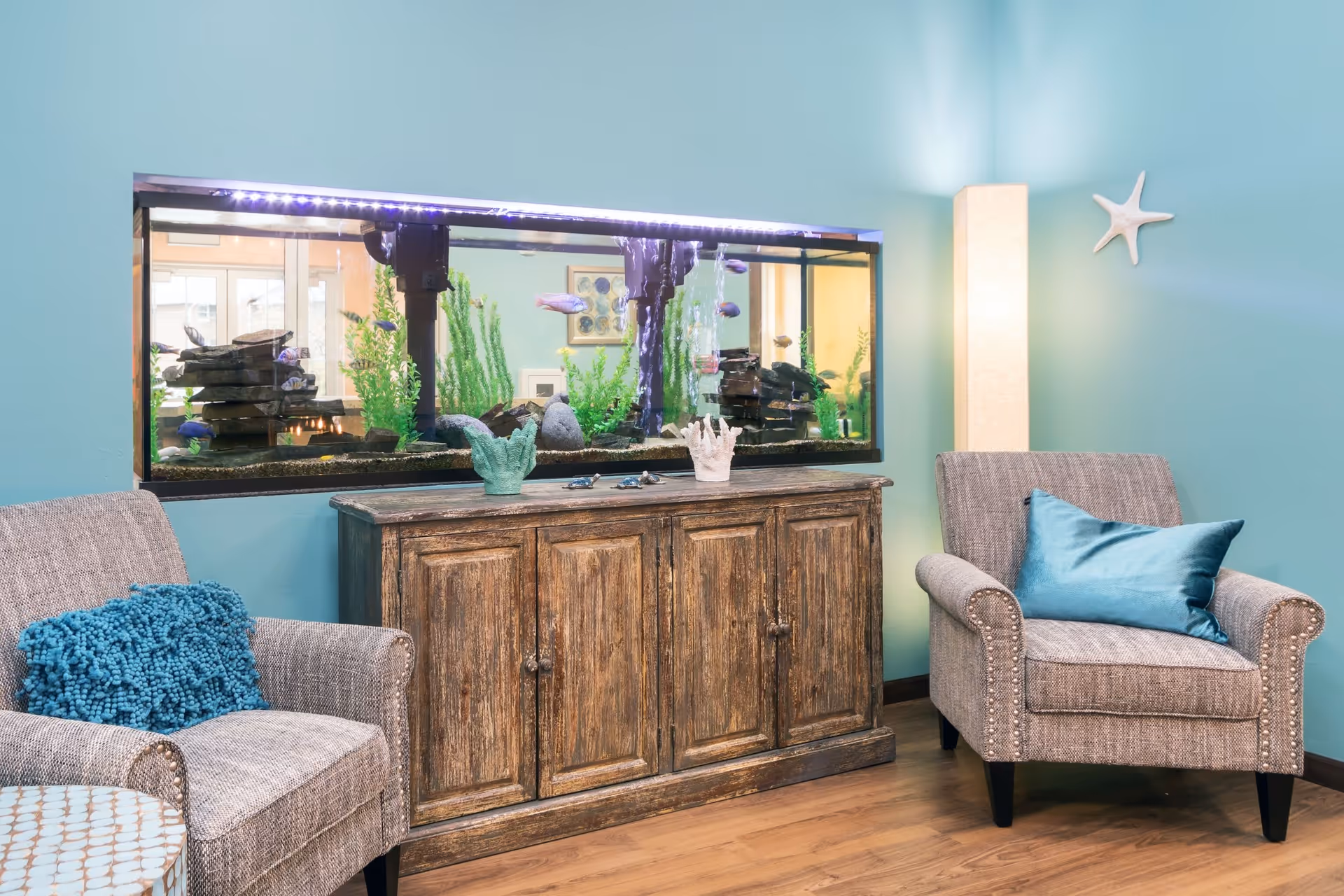 A cozy seating area with two gray upholstered armchairs, each with a blue cushion, flanking a rustic wooden cabinet. Above the cabinet is a large built-in aquarium filled with fish and aquatic plants. The walls are painted light blue, with a tall floor lamp and a white starfish decoration on the wall.