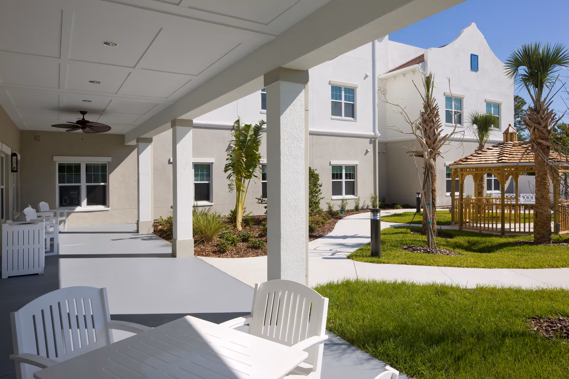 Outdoor covered patio area with white chairs and tables, adjacent to a garden with green grass, palm trees, and a wooden gazebo. The building exterior is light-colored with multiple windows under a clear blue sky.