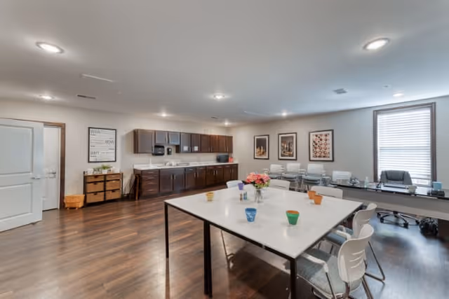 A spacious, well-lit room with a large white table in the center surrounded by white chairs. On the table are colorful cups and a vase with pink flowers. Along one wall, there are dark wooden cabinets with a countertop, a sink, and a microwave. The room has wooden flooring, framed artwork on the walls, and a window with blinds letting in natural light. There is also a desk with an office chair near the window.