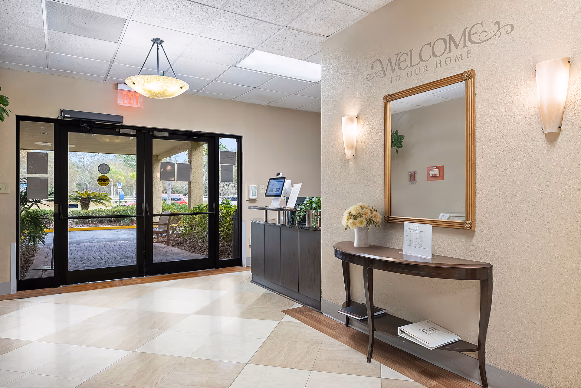 Lobby entrance with glass double doors, a reception desk, a mirror and console table under a 'Welcome to Our Home' sign.