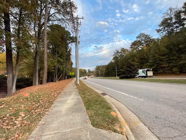 A sidewalk running alongside a road with a few cars and a truck driving on it. Trees line the left side of the sidewalk, and there is a grassy strip between the sidewalk and the road. The sky is partly cloudy with patches of blue visible.