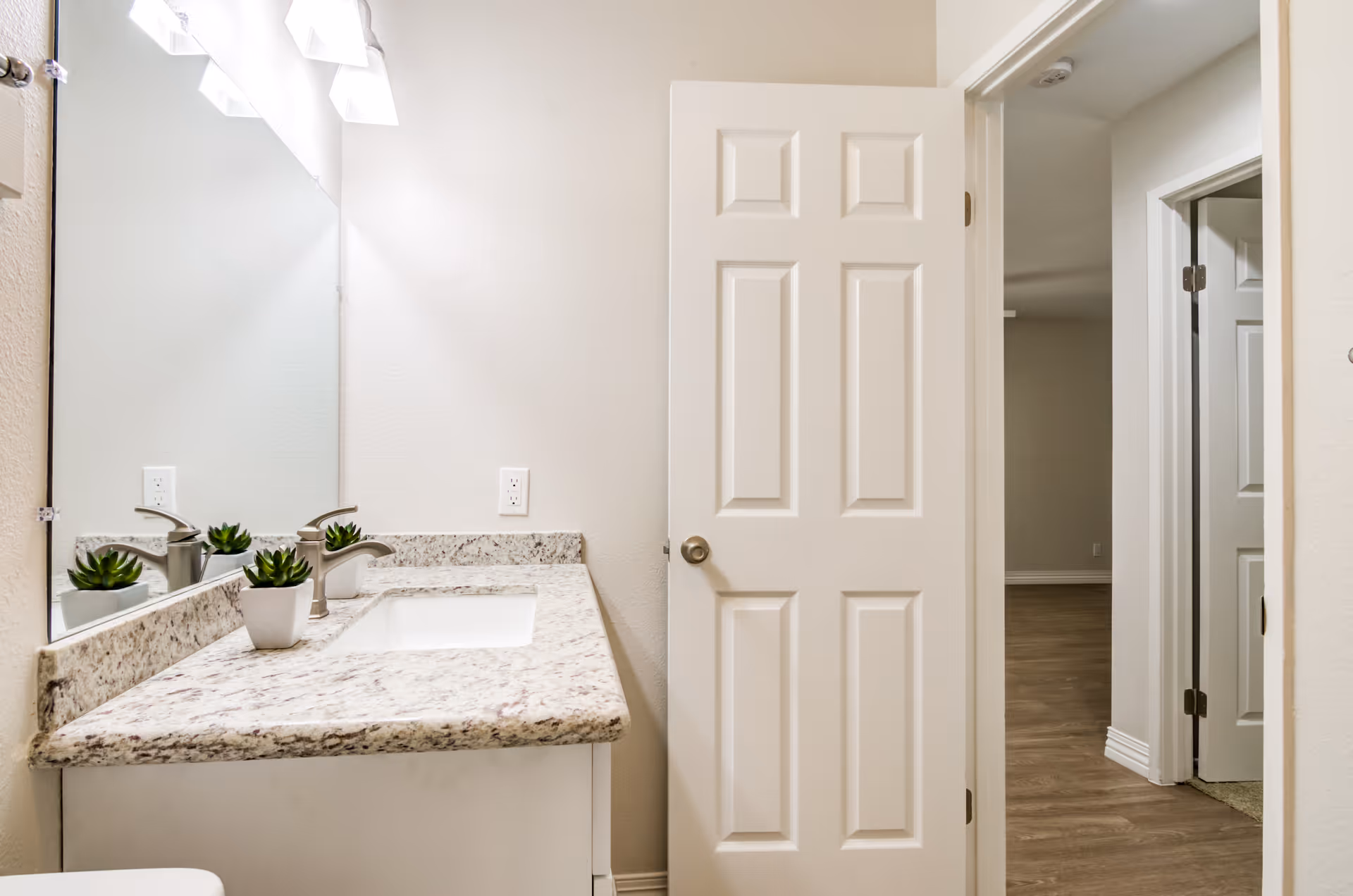 A bathroom interior featuring a granite countertop with a built-in sink, a modern faucet, and a small potted plant. A large mirror is mounted above the countertop with three light fixtures above it. The bathroom door is open, showing a hallway with wooden flooring and additional doors.