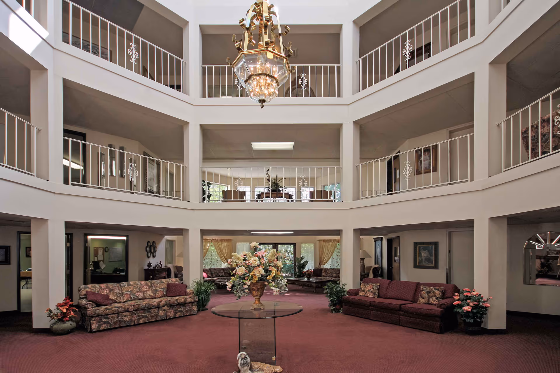 Interior view of a senior living facility common area with a high ceiling and two levels of white railings. The room features floral and solid-colored sofas, a glass table with a large flower arrangement in the center, potted plants, and a large chandelier hanging from the ceiling.
