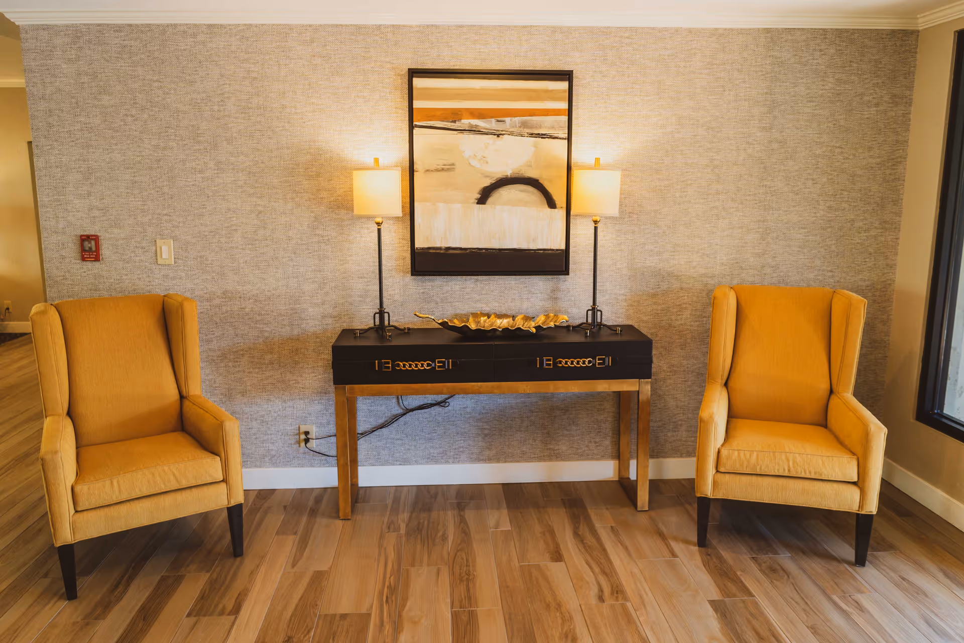 A cozy interior seating area with two mustard yellow armchairs placed on either side of a black and gold console table. The table holds a decorative gold bowl and two tall lamps with beige shades. Above the table hangs an abstract painting with neutral tones. The floor is wooden, and the walls have a textured beige wallpaper.