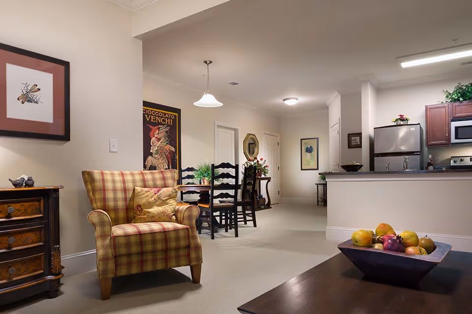 Interior view of a senior living facility showing a cozy living area with a plaid armchair and a wooden side table with decorative items. In the background, there is a dining area with a table and chairs, and a kitchen with stainless steel appliances and wooden cabinets. The space is well-lit with ceiling lights and decorated with framed artwork and plants.