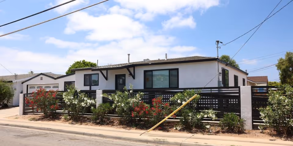 Exterior view of a single-story assisted living facility building with white walls and a dark roof, surrounded by a black fence with white pillars and flowering bushes in front. The sky is partly cloudy.