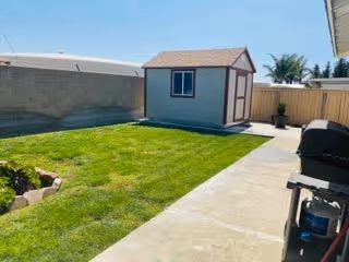 Small backyard with a green lawn, concrete walkway leading to a storage shed, fenced perimeter, and a grill on the right.