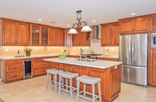 A spacious kitchen with wooden cabinets, a large island with a white countertop, three cushioned stools, stainless steel refrigerator, built-in microwave, and a wine cooler. The kitchen is well-lit with recessed lighting and a decorative hanging light fixture above the island.