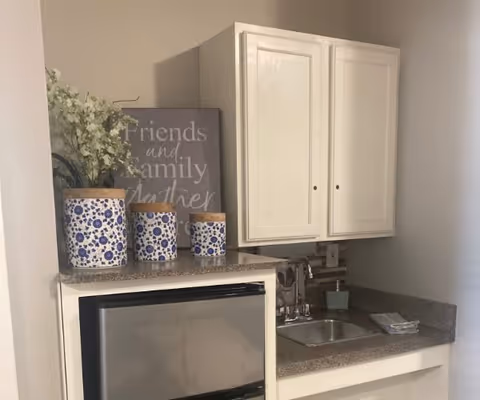 A small kitchenette area with a mini refrigerator, a countertop with a sink, and white cabinets above. On the countertop, there are three decorative blue and white canisters with wooden lids and a vase with white flowers. A framed sign with the words 'Friends and Family Gather' is placed behind the canisters.