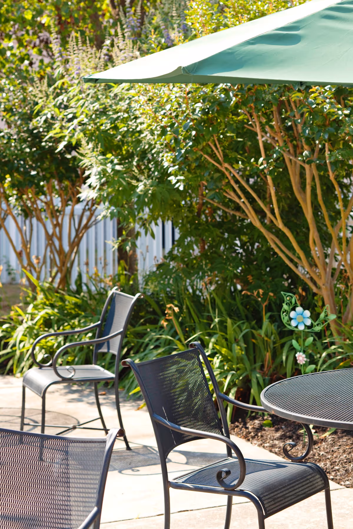 Outdoor patio with metal chairs and a round table under a green umbrella next to shrubs and plants.