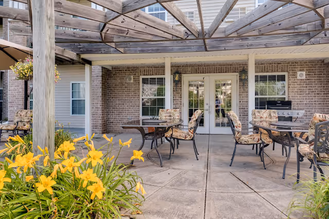 Outdoor patio area with a wooden pergola overhead, several tables with cushioned chairs arranged on a concrete floor, yellow flowers in the foreground, and a brick building with glass double doors and windows in the background.