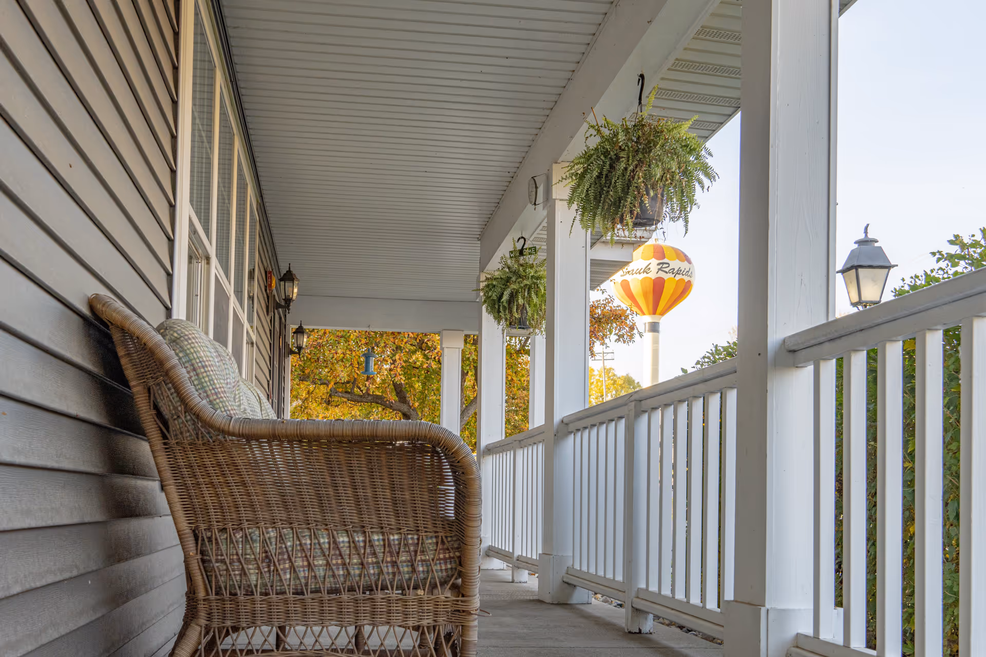 A covered porch with white railings and ceiling, featuring a wicker chair with cushions on the left side. Hanging plants are suspended from the ceiling, and a colorful water tower with the text 'South Rapids' is visible in the background among trees with autumn foliage.